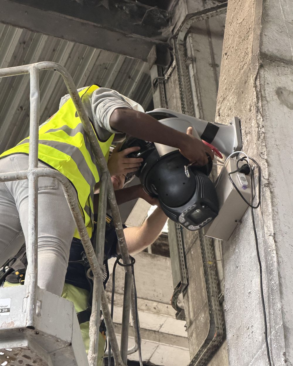 Pose d'une caméra dôme et de son boîtier de traitement sur un pilier en béton, depuis une nacelle, dans une unité de traitement des déchets.